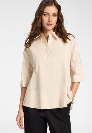 Kobieta o falowanych brązowych włosach, ubrana w jasnobeżową, luźną koszulę z rękawami do łokcia i czarne spodnie, stojąca z jedną ręką w kieszeni.