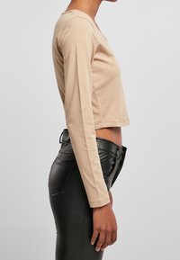 Beige-langärmliges Oberteil mit Rundhalsausschnitt, kombiniert mit hochgeschnittenen schwarzen Kunstlederhosen, die eine verkürzte Passform betonen.