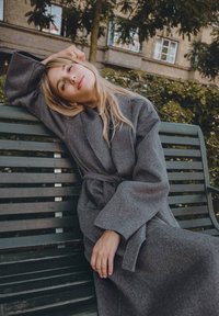 Szary wełniany szlafrok z paskiem w talii, szerokimi rękawami i miękką teksturą, na tle zielonej ławki i liściastego krajobrazu.