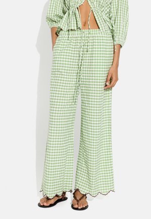 Osoba nosząca jasnozielone i białe szerokie spodnie w kratę z regulowanym pasem i brązowe plecione sandały, ręce w kieszeniach.
