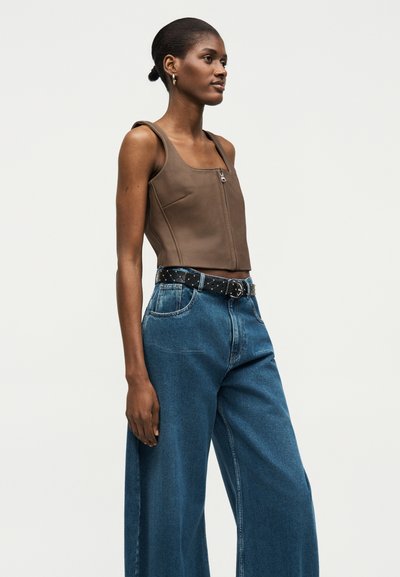 Γυναίκα στέκεται φορώντας καφέ αμάνικη μπλούζα με φερμουάρ μπροστά, μπλε τζιν παντελόνι με φαρδιά πόδια και μαύρη ζώνη με ασημένια καρφιά σε απλό φόντο.