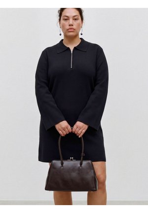 Femme portant une robe noire à manches longues avec une fermeture éclair à l'avant, tenant un sac à main marron foncé des deux mains devant son corps.