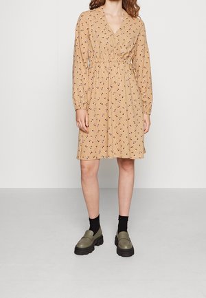 Kobieta w beżowej, kwiecistej sukience z długim rękawem sięgającej do kolan, do której dobrano czarne skarpetki i masywne oliwkowozielone mokasyny.