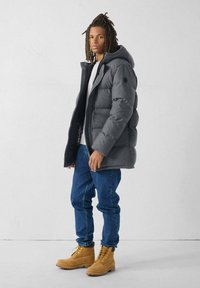 Giubbotto grigio imbottito con cappuccio, caratterizzato da un design trapuntato spesso, chiusura con zip e logo nero rotondo. Abbinato a jeans blu e stivali color cuoio.