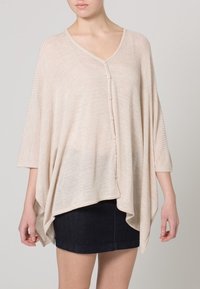 Donna che indossa un ampio poncho beige con bottoni e maniche a tre quarti, abbinato a una mini gonna in jeans scuri.