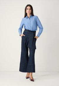 Blaues Oberhemd mit langenÄrmeln, kombiniert mit hoch taillierten, weit geschnittenen Jeanshosen aus dunklem Denim mit einem gebundenen Gürtel. Lackleder-Fersen.