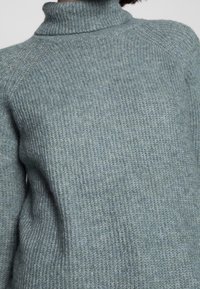 Světlomodrý pletený svetr s vysokým límcem, s žebrovanou texturou a raglánovými rukávy, ukazující měkký a teplý materiál.