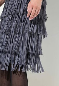 Robe frangée grise avec des bandes текстurées superposées. Main reposant sur la robe. Collants noirs partiellement visibles. Fini en tissu lisse.