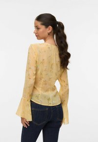 Blouse jaune à imprimé floral avec de longues manches évasées ; tissu léger et transparent ; détails de couture au dos ; portée avec un jean noir.