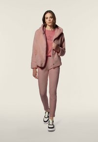 Giacca a collo alto rosa con cappuccio e pantaloni abbinati; texture morbida, spalle scese e polsini a costine; abbinata a sneaker nere e bianche.