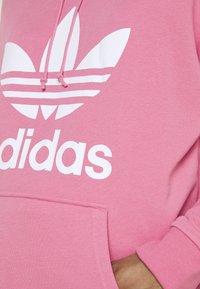 Nära bild av en rosa Adidas huvtröja med vit trefoil-logotyp och känguruficka, hand delvis inuti fickan på höger sida.