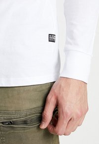 Vit långärmad T-shirt med en liten svart G-Star Raw-etikett. T-shirten har en figurnära design och en mjuk bomullstextur. Olivgröna byxor med en ficka som stängs med dragkedja.