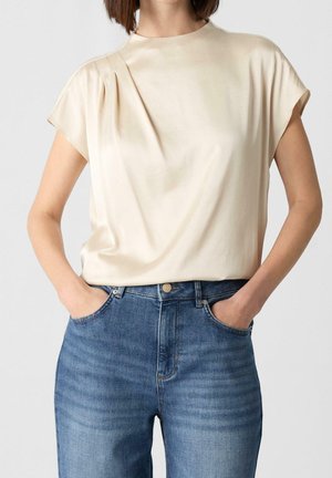 Kvinde iført en cremefarvet satinbluse med korte ærmer, stukket ned i højtaljede blå jeans med hænderne i lommerne.