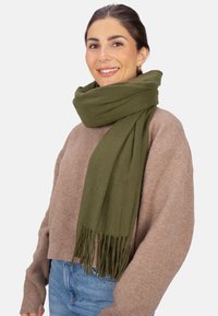 Écharpe en laine vert olive à franges, enroulée autour du cou, associée à un pull beige clair et un jean en denim bleu.