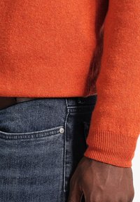Orange stickad tröja med ribbad mudd, parat med mörka denimjeans. Texturen är mjuk och färgerna kontrasterar livligt.