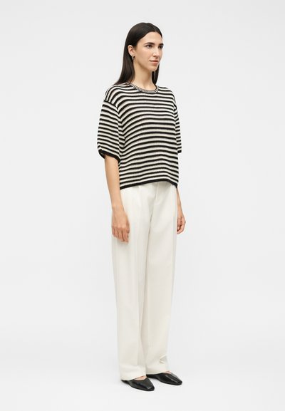 Γυναίκα στέκεται φορώντας μαύρο και λευκό ριγέ μπλουζάκι με κοντά μανίκια, λευκό παντελόνι με φαρδιά μπατζάκια και μαύρες μυτερές μπαλαρίνες σε απλό φόντο.