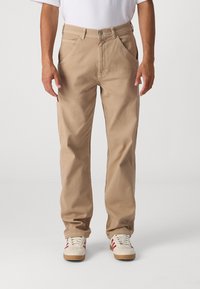 Beige cotton pants with a loose fit, front pockets, and side cargo pocket. Paired with white sneakers featuring red stripes.