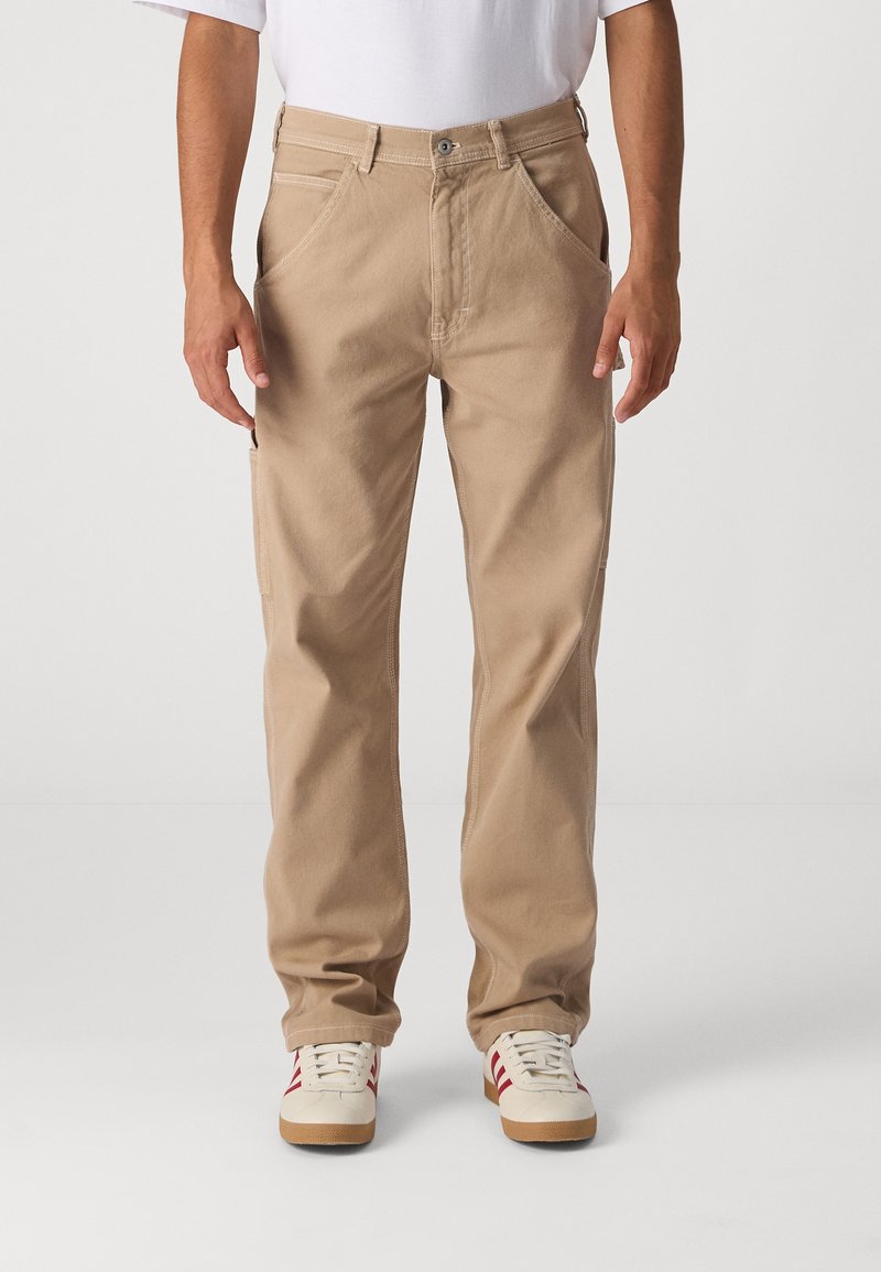 Beige cotton pants with a loose fit, front pockets, and side cargo pocket. Paired with white sneakers featuring red stripes.