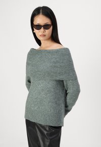 Zeleni pulover iz volnenega mešanice z off-the-shoulder dizajnom, rebrastim ovratnikom in teksturiranim telesom, v kombinaciji z sijočimi črnimi usnjenimi hlačami.