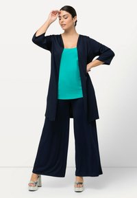 Duster bleu marine à manches longues sur un débardeur teal. Un pantalon noir à jambes larges et des talons plateformes argentés complètent la tenue.