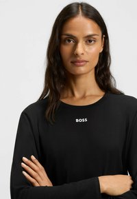 Musta pitkähihaine t-paita, joka on valmistettu sileästä kankaasta. Paidassa on valkoinen "BOSS" -logo keskellä rintaa, pyöreä pääntie ja rento istuvuus.