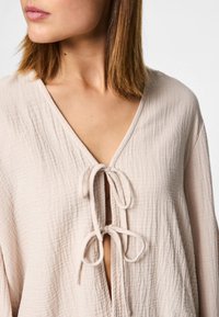 Light beige, textured top with a V-neck and tied front closure. Features soft fabric with a slightly crinkled appearance.
