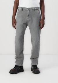 Pantalones de algodón gris con diseño de pierna recta, con cierre de botón y cremallera. Combinados con calzado negro grueso.