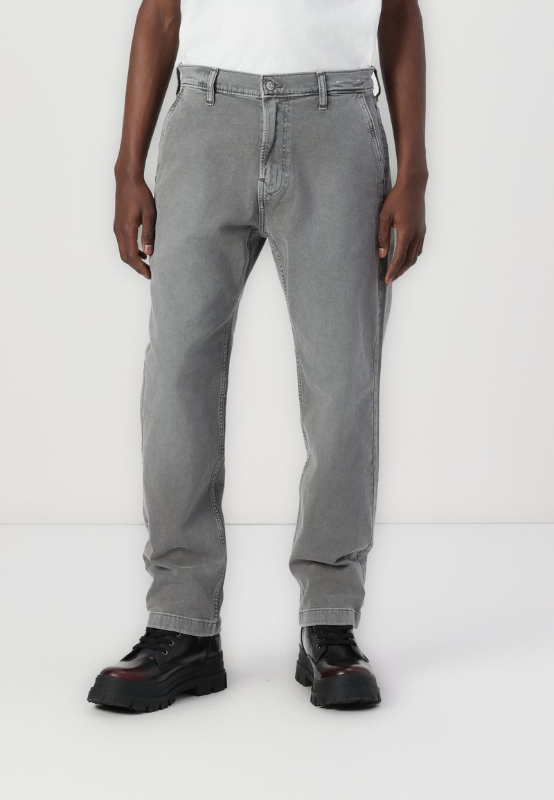 Pantalones de algodón gris con diseño de pierna recta, con cierre de botón y cremallera. Combinados con calzado negro grueso.