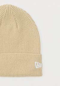 Bež pleteni vuneni šešir s presavijenim rubom i malim bijelim izvezenim logotipom na manžeti, prikazan na bijeloj pozadini.