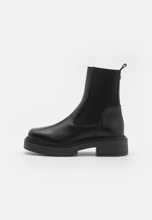 Stivale alla caviglia nero con tomaia in pelle liscia e pannelli elastici laterali. Presenta una suola spessa con finitura texturizzata e una linguetta.