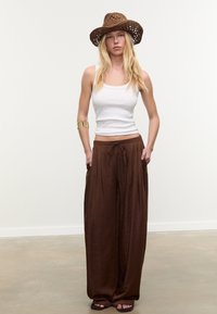 Pantaloni larghi marroni con vita regolabile e tasche laterali, abbinati a una canotta bianca e a un cappello di paglia intrecciato marrone. Sandali semplici.