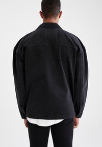 Musta denimhousutakki, jossa on rento leikkaus, näkyvät saumat ja takana logo. Yhdistetty valkoisen paidan kanssa, joka on kerroksittain alla.