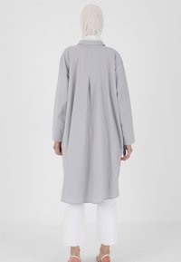 Longue tunique grise avec un col, fermeture à boutons et un pli à l'arrière. Associée à un pantalon blanc à jambe large et des sandales simples.