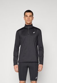 Svart treningsoverdel med lange ermer, høy krage og halv glidelås, laget av glatt stoff. Brukes med svarte tettsittende shorts.