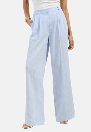 Kobieta nosząca jasnoniebieskie szerokie spodnie z plisami, w połączeniu z białym topem i złotą bransoletką, stojąca z jedną ręką w kieszeni.