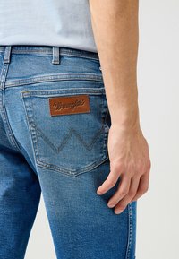 Modra kavbojkasta z malim usnjenim logom Wrangler na zadnjem žepu in kontrastnim šivom; slim-fit dizajn, gladka tekstura.