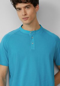 Turkusowa koszulka henley z krótkim rękawem, z okrągłym kołnierzykiem, trzyguzikowym zapięciem i gładką, lekką teksturą materiału.