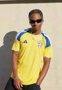 Maglia sportiva gialla con accenti blu, con l'emblema nazionale svedese. Il modello indossa occhiali da sole neri e gioielli d'argento.