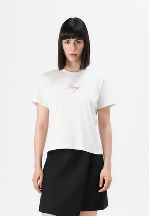 Woman with straight dark hair wearing a white Hugo t-shirt and a black asymmetrical skirt, standing against a plain background.