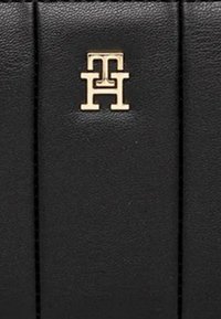 Borsa a mano in pelle nera con superficie strutturata, dettagli di cuciture verticali e un logo dorato con le iniziali "TH" ben visibili.