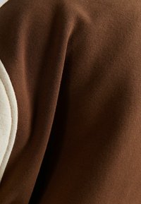 Brun sweatshirttyg med en mjuk textur, som har en kontrasterande ljus accent på axeln, vilket framhäver designens enkelhet.