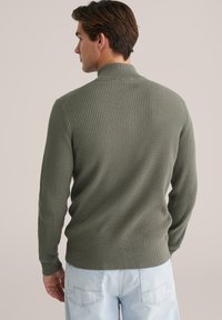 Man met zijn rug naar de camera, gekleed in een ribbelige olijfgroene trui met een hoge kraag en lichtblauwe spijkerbroek, tegen een effen achtergrond.