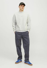 Ung mand iført en lys grå sweatshirt, mørke cargo bukser, blå sneakers og en sort kasket, står med hænderne i lommerne mod en hvid baggrund.