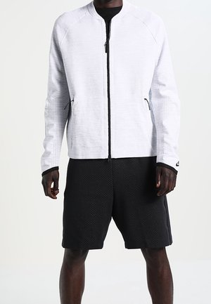 Homme portant une veste blanche zippée avec des manches texturées et un short de sport noir, se tenant devant un fond uni.
