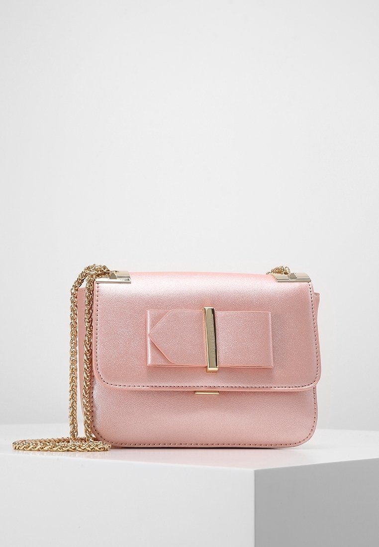 Růžová metalická kabelka s předním klopou, detailem mašle, zlatými kovovými prvky a řetízkem na nošení. Hladká textura a obdélníkový tvar.