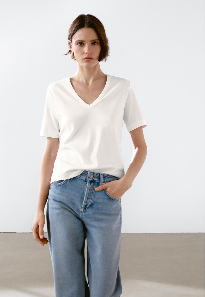 Biała koszulka typu V-neck z miękkiego materiału, z krótkimi rękawami, w połączeniu z jasnoniebieskimi spodniami typu wide-leg. Modelka ma jedną rękę w kieszeni, na prostej tle.