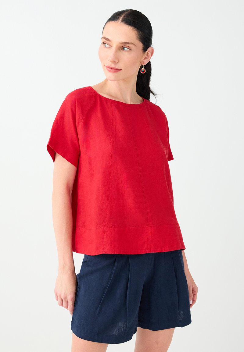 Femme aux cheveux foncés attachés en queue de cheval, portant un haut rouge à manches courtes et un short bleu marine, debout devant un fond blanc uni.