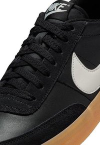 Zapatilla negra con materiales de ante y cuero, que presenta cordones planos negros, un logotipo de Nike en blanco y una suela de goma color miel.