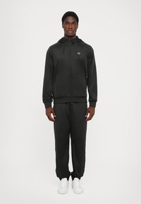 Svart träningsdräkt i mjukt tyg, bestående av en zip-up hoodie och byxor med elastisk midja. Inkluderar en liten logotyp och framfickor.