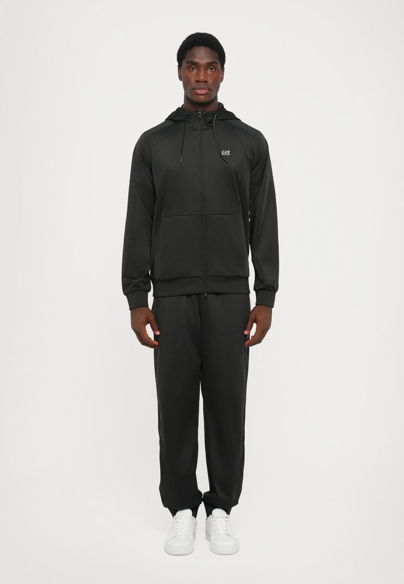 Svart träningsdräkt i mjukt tyg, bestående av en zip-up hoodie och byxor med elastisk midja. Inkluderar en liten logotyp och framfickor.